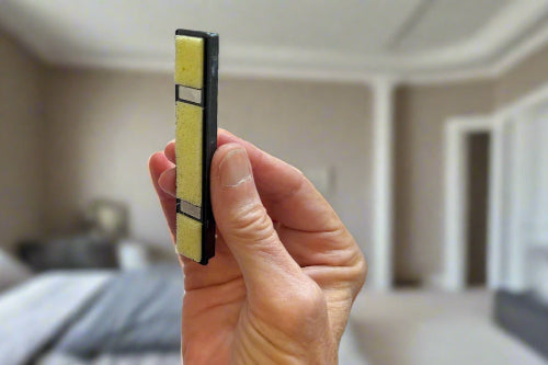 Hand holding a Hinge Husher lubricant cartridge in front of a bedroom scene